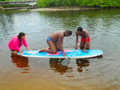 One athlete kneeling on a stand up paddle board in the water. One coach standing in the water holding the back of the paddle board. Another coach standing in the water, leaning on the paddle board, holding the paddle. Green woods and bridge in the background. 