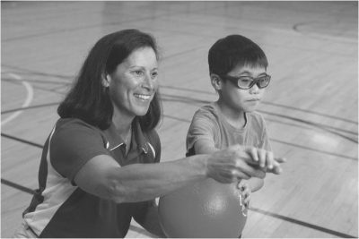 Lauren using hand-over-hand guidance with an young athlete touching a ball in greyscale. Athlete has short brown hair and wearing glasses holding Lauren's hand. Lauren smiling and helping athlete wearing a polo with short brown hair at her shoulders.