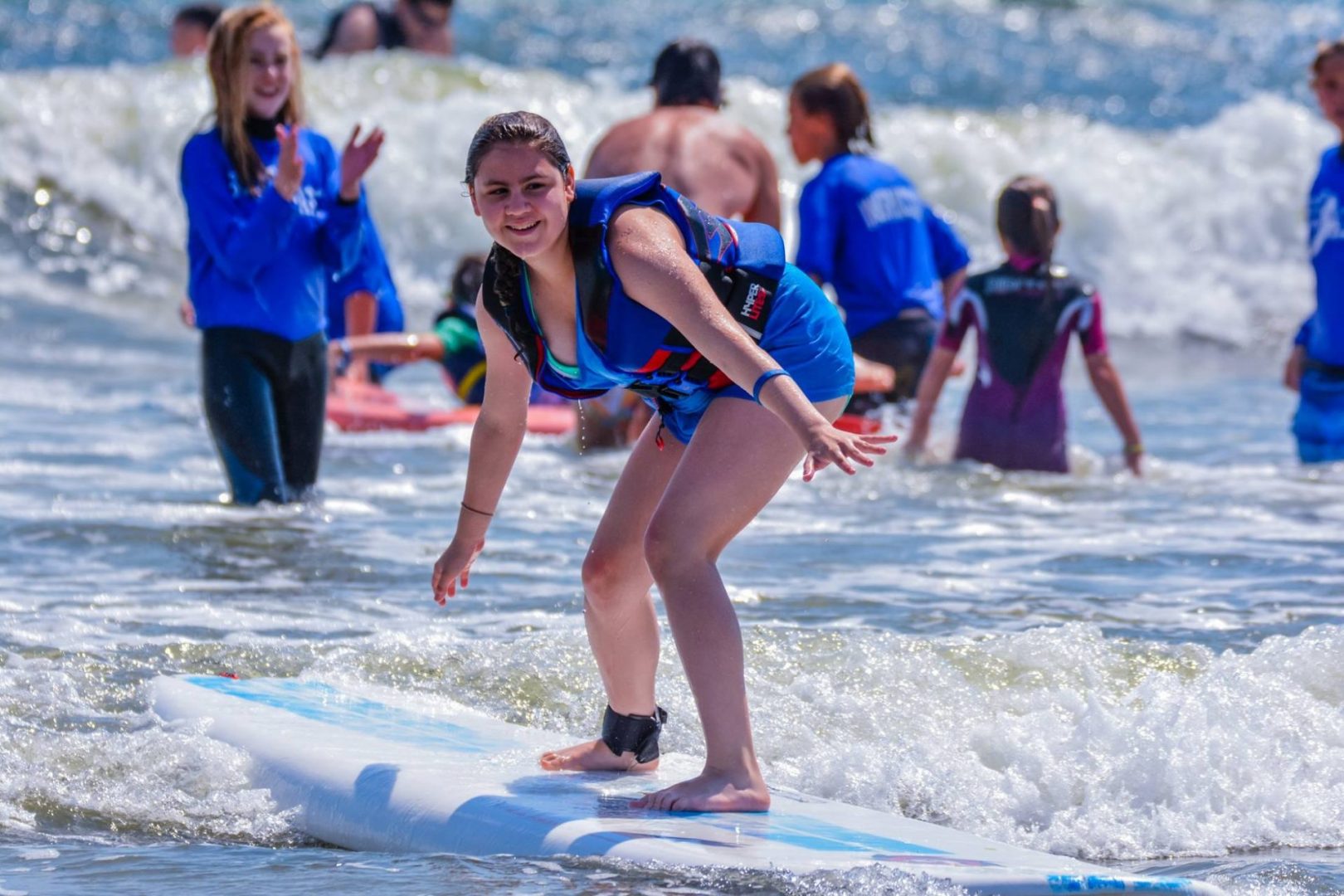 Lots of athletes and coaches in the water with large waves. Photo is focused on one athlete who is standing on her surfboard. She is wearing a life vest. Her knees are slightly bent and she is riding towards the shore with the wave. A coach in the background is clapping for her.