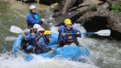 Athletes and coaches in a raft navigating rapids for white water rafting. People are smiling and looking shocked trying to navigate the rapids.