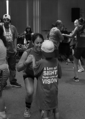 Maria bending down and dancing with an athlete. Athlete is wearing an oversized shirt and baseball cap and Maria is holding both of the athlete's hands and smiling. Photo in greyscale.
