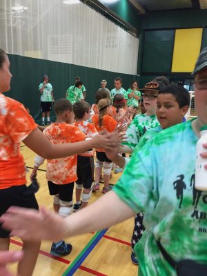 Athletes and coaches from orange and blue teams lined up high-fiving each other in the Brockport gym. Wooden floor with colorful lines and green and yellow mats on the walls. A green net with mesh top is pulled down separating the gym. 