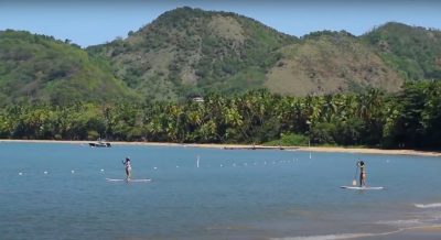 Landscape photo of Puerto Rico. Large blue ocean with sandy beach and large green mountains in the background. Two people stand up paddle boarding in the water.