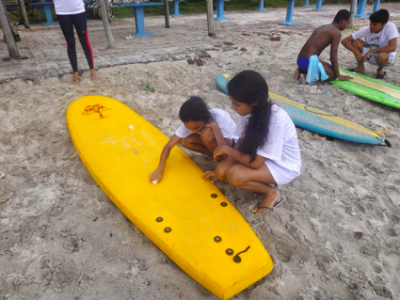 Athlete and coach kneeling in the sand. Athlete is tactile feeling the yellow paddle board with their hand with coach next to them. Two more paddle boards in the background with another athlete and coach knelt in the sand. Athlete is tactile feeling the paddle board with their hands. 