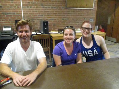 Griffin, Lindsay, and Martha sitting in a line at a table smiling at the camera. A brick wall with desks and computer equipment is behind them. 