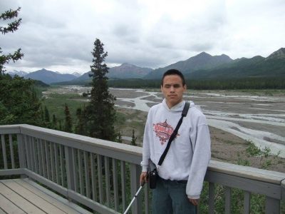 Paul posing for a photo while holding his cane. He is standing on a wooden deck with large mountains far in the distance behind him. 