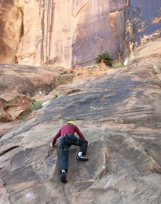 Paul rock climbing up a large rock face. He is wearing a harness and helmet. He is reaching one leg up and has both hands pressed against the rock as he climbs higher. 