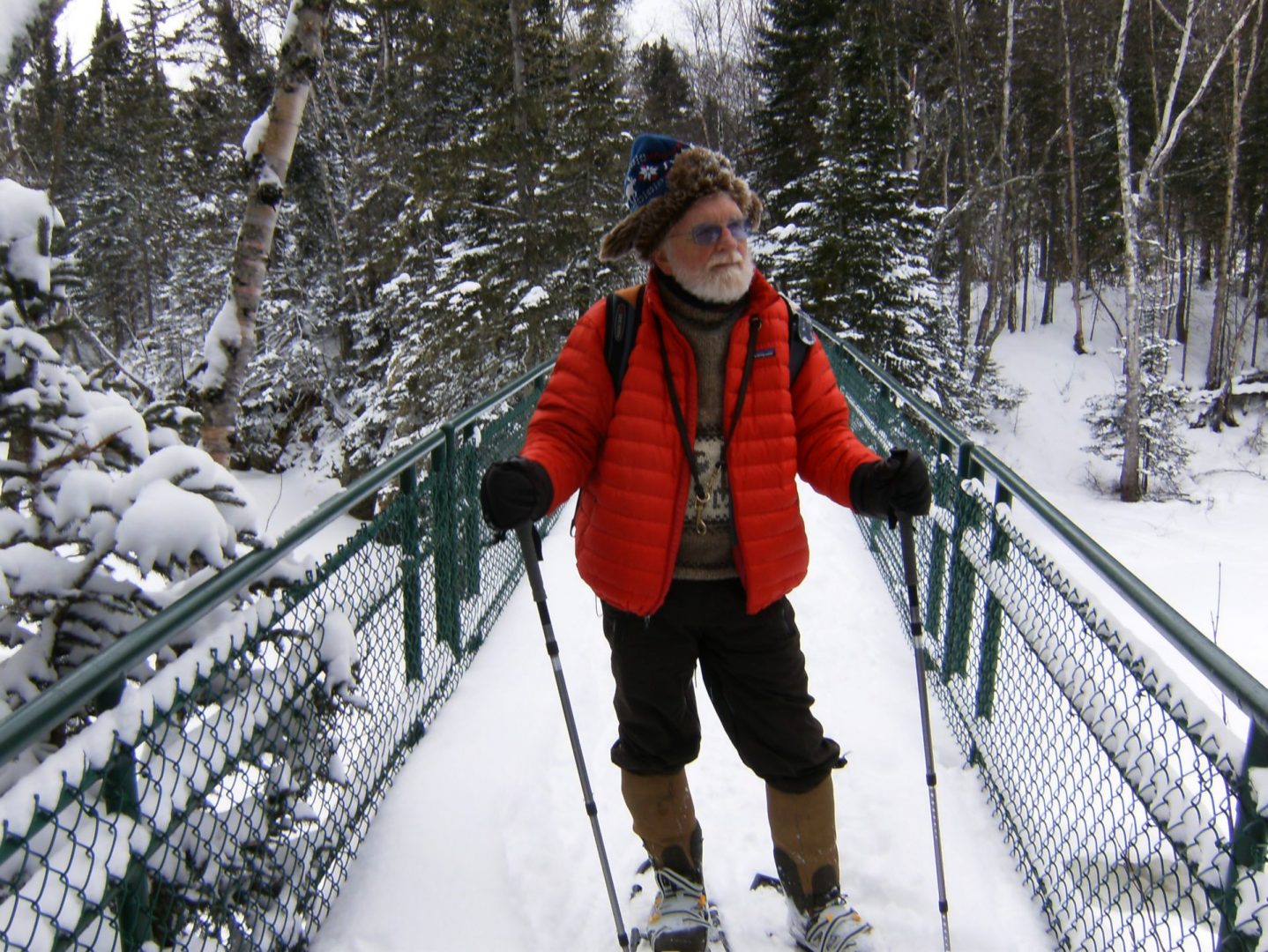 Paul posing for a photo in a snowy wooded area. He is standing on a bridge wearing his snowshoes holding walking poles in both hands. There is lots of snow in the background covering the trees and bridge.