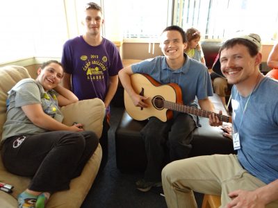 A photo of Paul and 3 peers. All 4 are smiling at the camera. Paul is holding a guitar in a position to being playing it. All four are sitting in a living room setting. 
