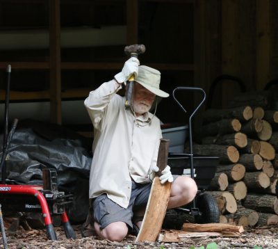 Paul is holding a hammer over his head as he is about to swing down to chop a piece of wood in half that he is balancing with his other hand. There is a pile of wood in the background outside. 