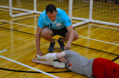 Coach Brian assisting an athlete with positioning their arms to stop the goalball while laying on their side. Goalball nets in the background. 