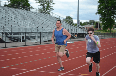 Coach Brian and athlete running on the track beside each other with bleachers in the background. Both individuals are pumping their arms and mid stride. 