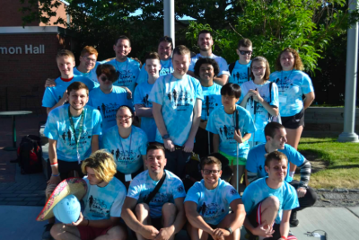 Group photo of Team Blue athletes and coaches smiling at the camera. Everyone is wearing their blue camp tshirts. Emerson Hall dormitory in background. 