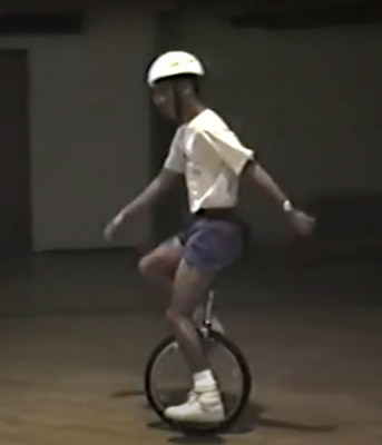 Eddie is inside a gymnasium riding a unicycle independently. He is wearing a helmet and one arm is in front of him and the other behind him as he balances on the bike. 