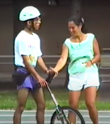 Eddie and Lauren standing on the outdoor track. Eddie is holding onto his unicycle while Lauren reaches for his hand. 