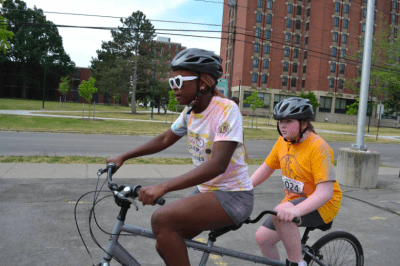 A coach on the front of a tandem bike with an athlete on the back. Both are wearing helmets. Large brick buildings in the background as they ride along the pavement. 