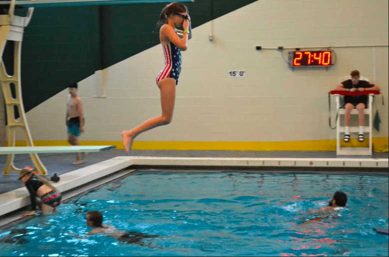 An athlete is hovering over the pool water after a jump of the diving board. She is wearing goggles and plugging her nose, preparing to land in the water. There are other athletes and coaches in the water. There is a lifeguard sitting in the lifeguard chair in the background.