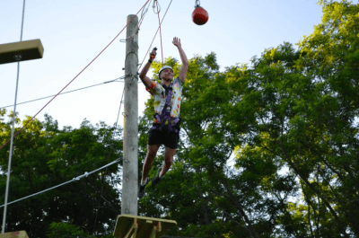 Athlete harnessed into a ropes course wearing a helmet. He is mid air after jumping off the wooden platform towards a red hanging ball. He is reaching his arms out in front of him to grab the hanging ball. The ropes from the course are attached to a large wooden pole. 