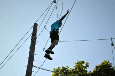 Athlete balancing on a tight rope while harnessed into the ropes course. He is wearing a helmet and sneaker. He has his arms out in the air beside him trying to keep his balance on the rope. The tops o the trees and blue sky are in the background. The ropes are attached to a large wooden pole. 