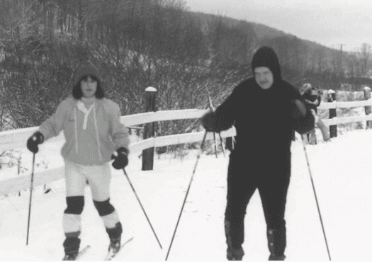 Black and white photo of Dr. Lieberman and colleague cross country skiing. Poles in both hands. Fence and woods in the background.