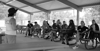 Lauren speaking in front of coaches underneath a pavilion. Lauren wearing a tank top, shorts, and bucket hat looking out to the rest of the crowd.