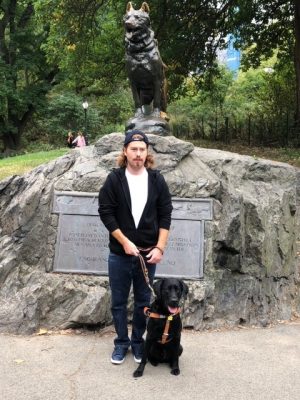 Griffin holding the leash of his service dog while the two stand in front of a statue of a dog in the background. 