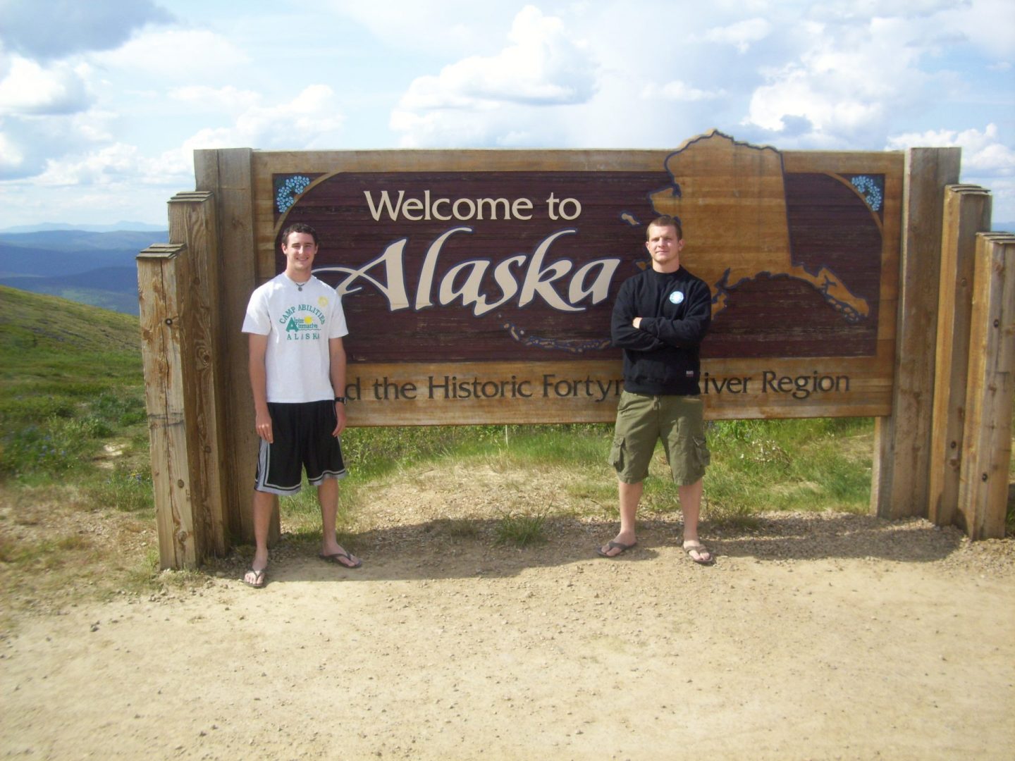 Matt and Justin posing in front of a sign that says "Welcome to Alaska". Blue sky and green mountains in the background.