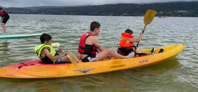 1 Coach and 2 athletes kayaking on the water. From the front to the back of the kayak, 1 athlete in orange lifejacket putting paddle into the water with a coach sitting in the middle with a red jacket and no paddle. The athlete in the back is wearing a bright green life jacket and holding paddle level out of the water.