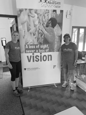 Photo of Val and Lauren Lieberman standing next to a Camp Abilities Suisse full-length poster. Photo in greyscale and both Val and Lauren are smiling wearing Camp Abilities Suisse shirts.