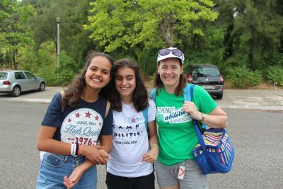 Ursula and two other individuals smiling at the camera with arms around each tother. Parked cars and green woods in the background. 