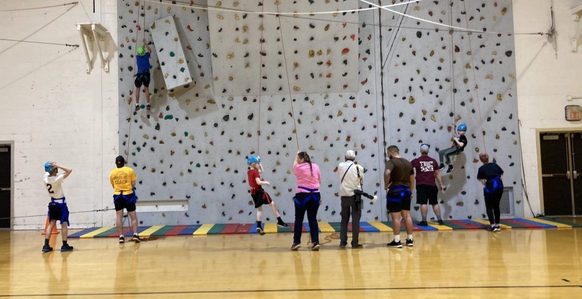 Photo of two athletes climbing the rock climbing wall in a gymnasium. Multiple people standing and watching or belaying the athletes in their harnesses.