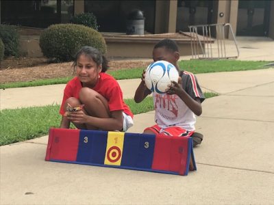 Raveena and another athlete are kneeled down on the sidewalk behind a score board on the ground. The other athlete is holding a soccer ball in front of his face. 