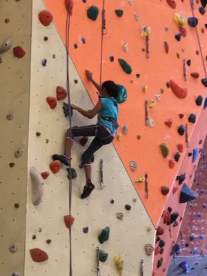 Athlete is climbing up an colorful indoor rock climbing wall. She is mid way up the wall and is harnessed in. She is using her arms and legs to move up the wall. 