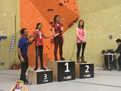 Raveena and two other athletes standing on podiums of different heights. Raveena is on podium number 3 representing her third place finish . The indoor rock climbing wall is in the background. The 1st place athlete is holding Raveena's hand and her coach is standing on the other side of her. 