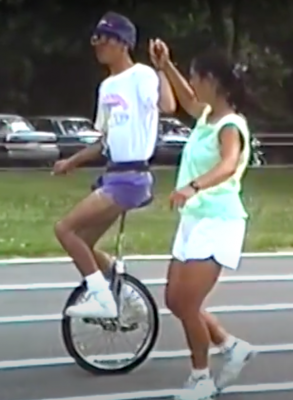 Eddie is riding a unicycle on an outdoor track. Lauren is standing next to him holding his hand while he balances on the bike. 