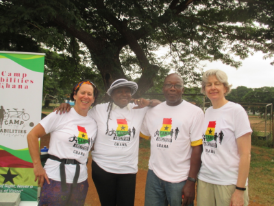 Lauren, Katrina and two other Camp Ghana volunteers smiling for a photo. Camp Abilities Ghana sign with the Camp Abilities Logo of a tandem bike and two silhouettes running, is behind them. They are all wearing their Camp Abilities Ghana camp tshirts with are white with red, yellow and green representing the Ghana flag. 