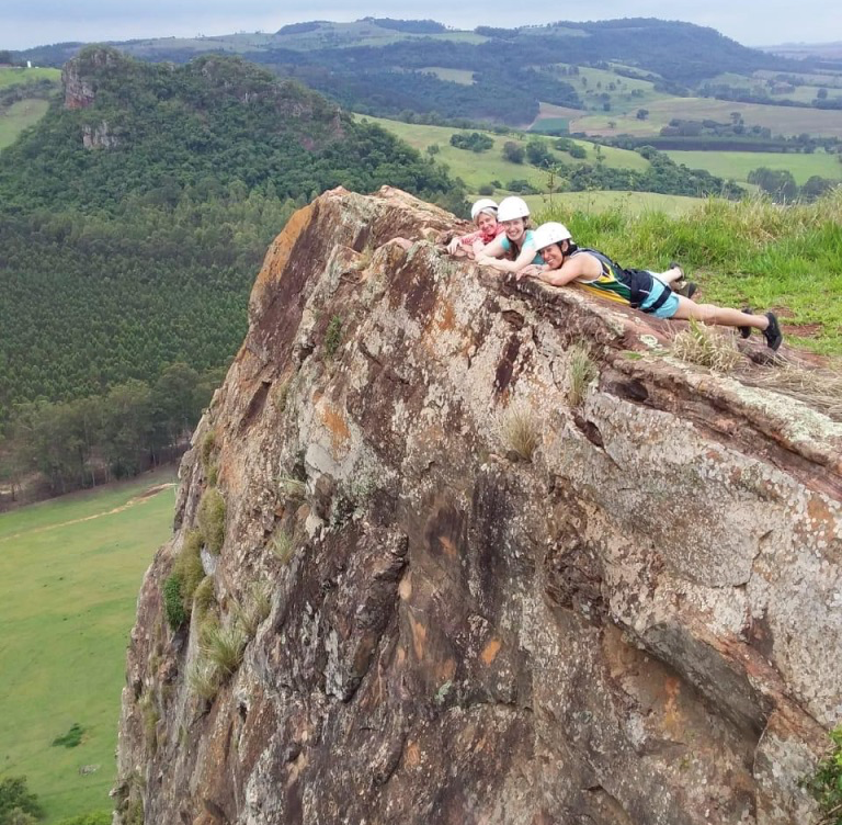 Lauren and two others laying on their stomachs at the top of a large rock. There are green mountains and trees extending far into the distance behind them. They are all wearing rock climbing gears including helmets.