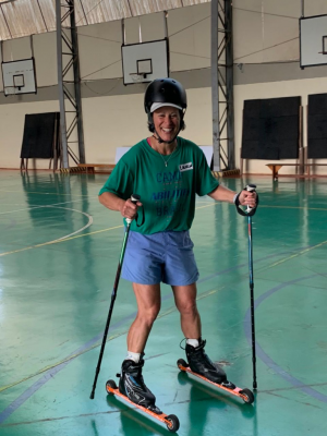 Lauren smiling big while holding ski poles and balancing on her roller skis. She is wearing a helmet and is indoors in a gymnasium with a green floor and 3 basketball nets in the background. 