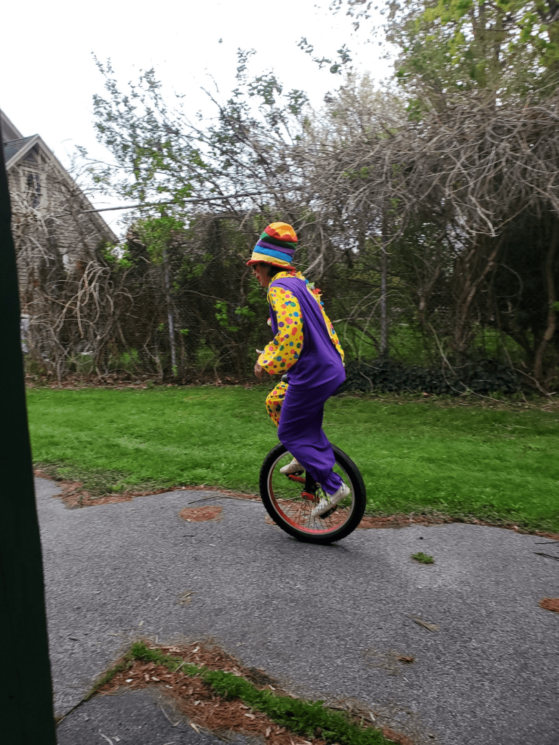 Dr. Lauren Lieberman wearing a colorful clown suit and hat while riding a unicycle on the pavement. Grass and trees are behind her in the background.