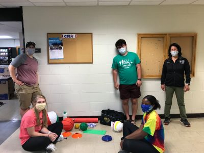Lauren and 4 other camp abilities graduate assistants and specialists wearing masks while sorting gear into bags to be mailed to camp abilities virtual athletes. Lots of equipment on the ground ready to be placed in a the gym bag that says Camp Abilities on it. 