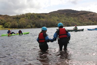 Athlete and coach walking into deeper water holding hands. Both are wearing helmets and life vests. Four individuals in kayaks and large green mountains in the background. 